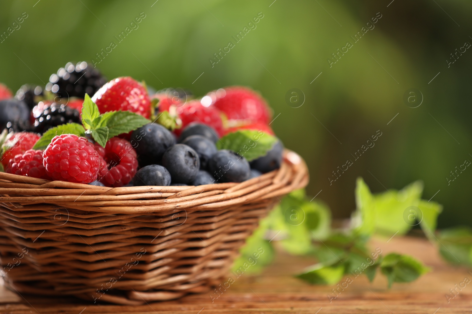 Photo of Wicker bowl with different fresh ripe berries and mint on wooden table outdoors, space for text