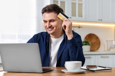 Photo of Happy man with credit card and laptop shopping online at wooden table in kitchen