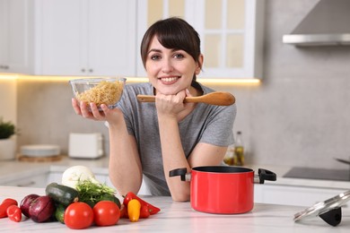 Happy young housewife with spoon and raw pasta at white marble table in kitchen. Cooking process