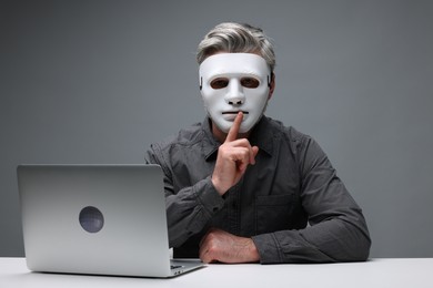 Photo of Man in mask showing hush gesture at white table with laptop against grey background