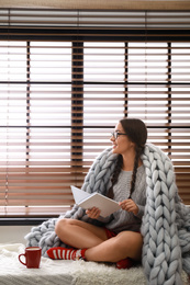 Happy young woman wrapped in knitted blanket with book near window at home. Lazy morning