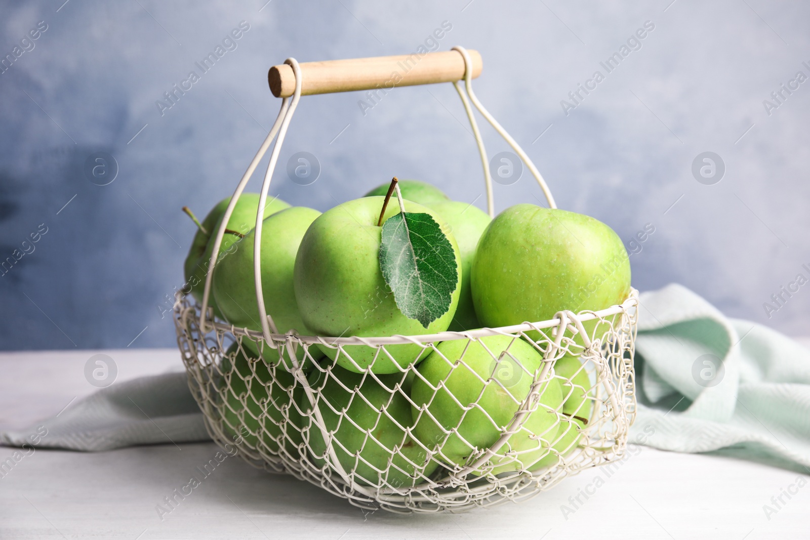 Photo of Metal basket of fresh ripe green apples on white wooden table against blue background