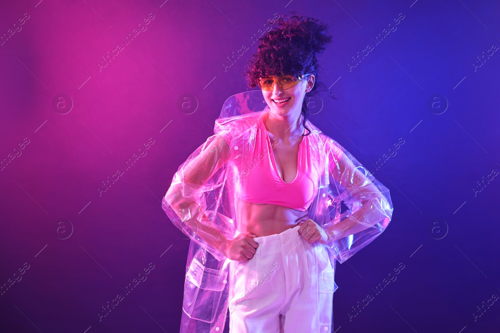 Photo of Beautiful young woman in transparent coat and sunglasses posing on color background in neon lights