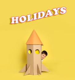 Image of School holidays. Cute little child playing with cardboard rocket near yellow wall