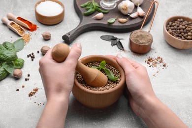 Woman mixing peppercorns and basil in mortar at light grey table, closeup