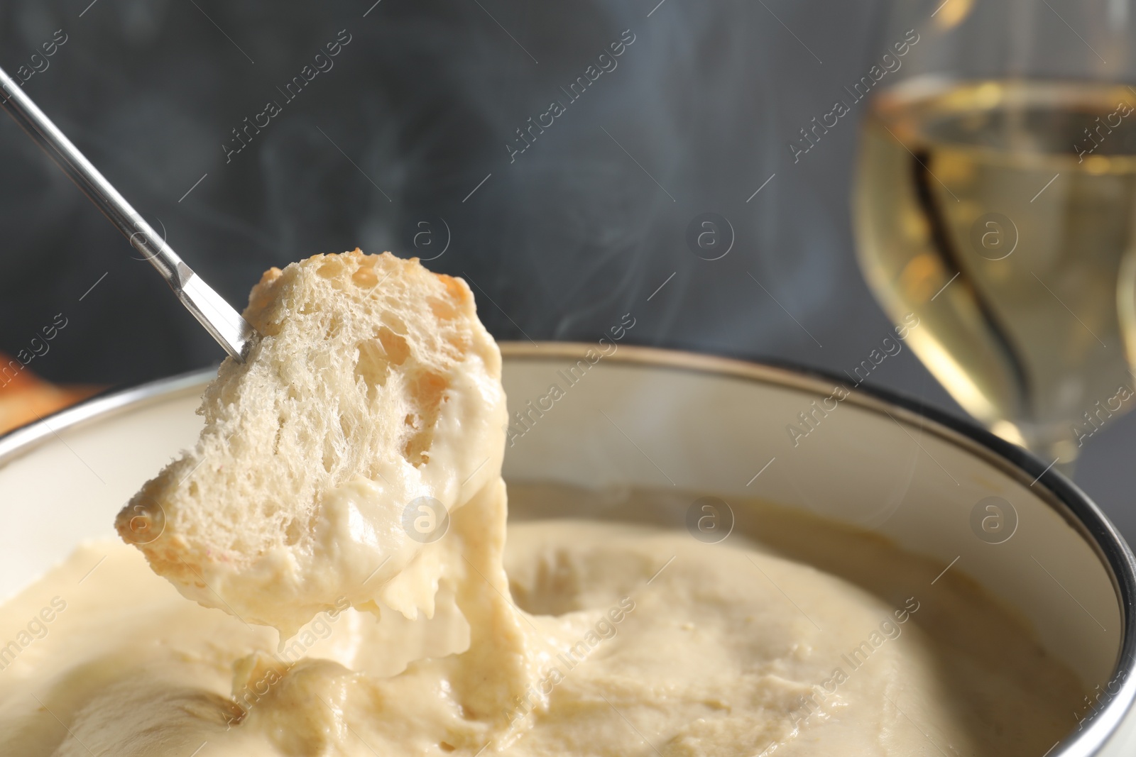 Photo of Dipping piece of bread into fondue pot with melted cheese on grey background, closeup