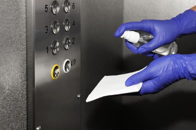 Photo of Woman with detergent spray and paper napkin cleaning elevator`s panel, closeup