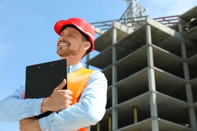 Photo of Professional engineer in safety equipment with clipboard at construction site. Space for text