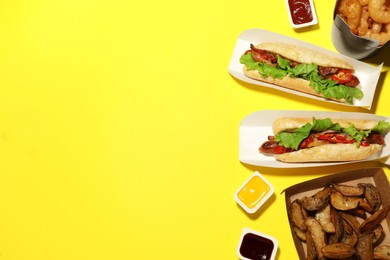 Photo of Tasty hot dogs, potato wedges, fried onion rings and different sauces on yellow background, flat lay with space for text. Fast food