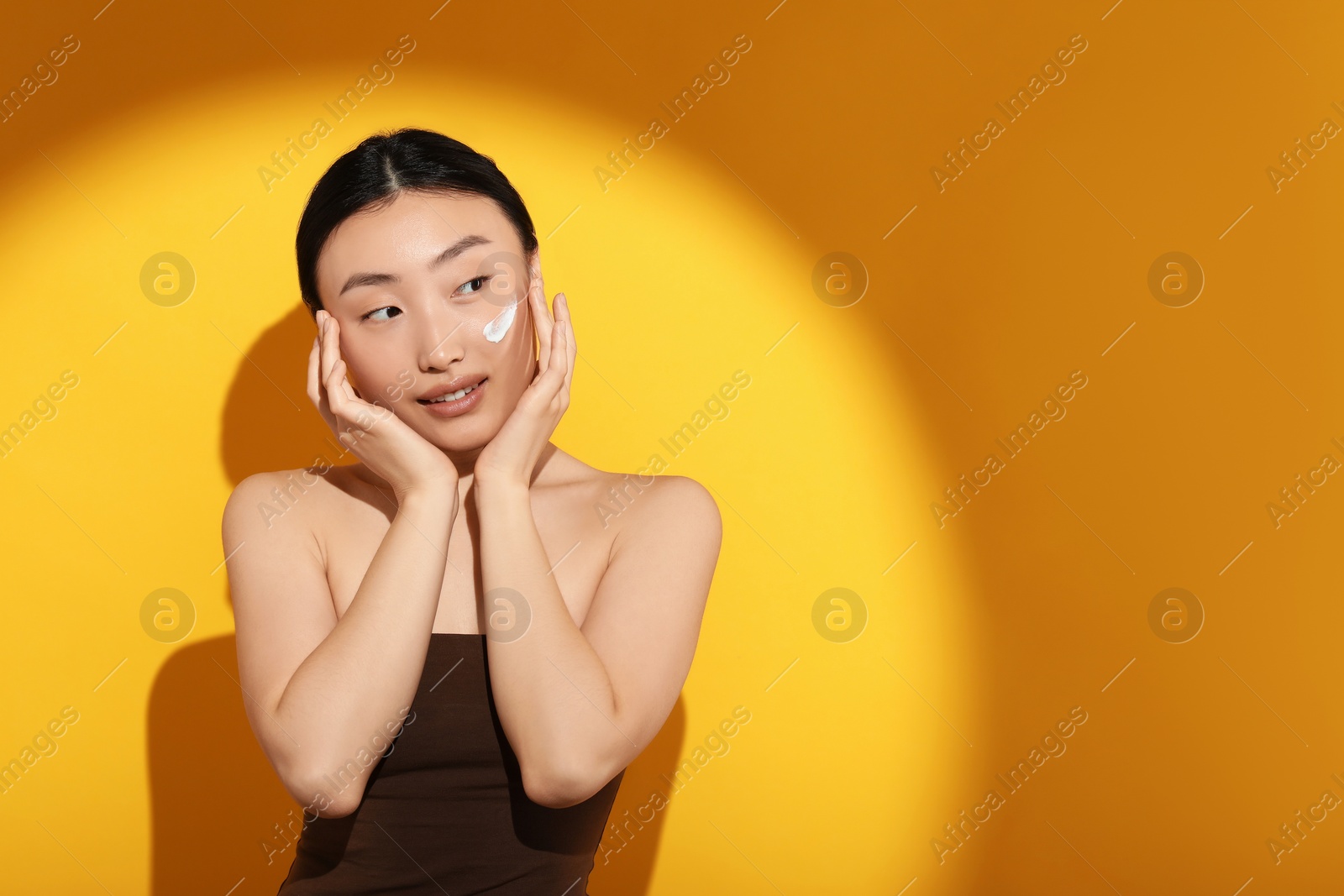 Photo of Beautiful young woman in sunlight with sun protection cream on her face against orange background, space for text
