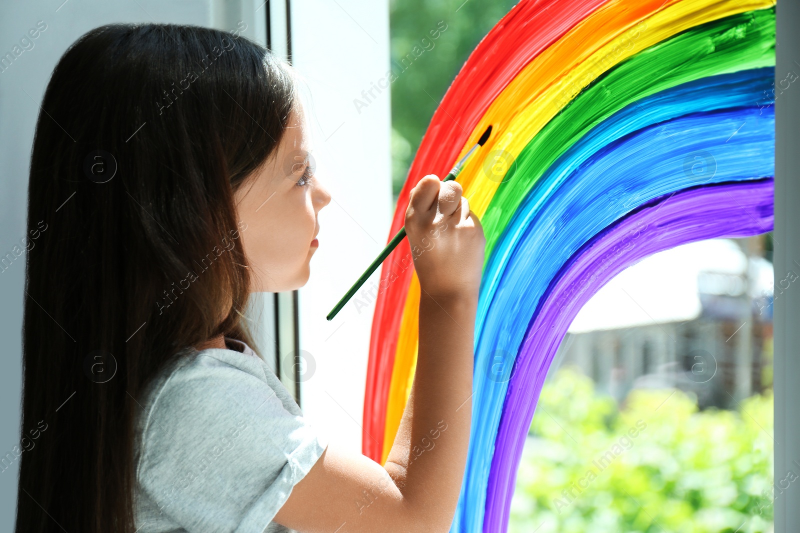 Photo of Little girl drawing rainbow on window indoors. Stay at home concept