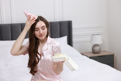 Beautiful young woman in party hat holding her Birthday cake on bed at home