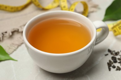 Photo of Cup of herbal diet tea and measuring tape on light marble table, closeup. Weight loss concept