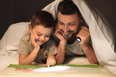 Photo of Father and son with flashlight reading book under blanket at home