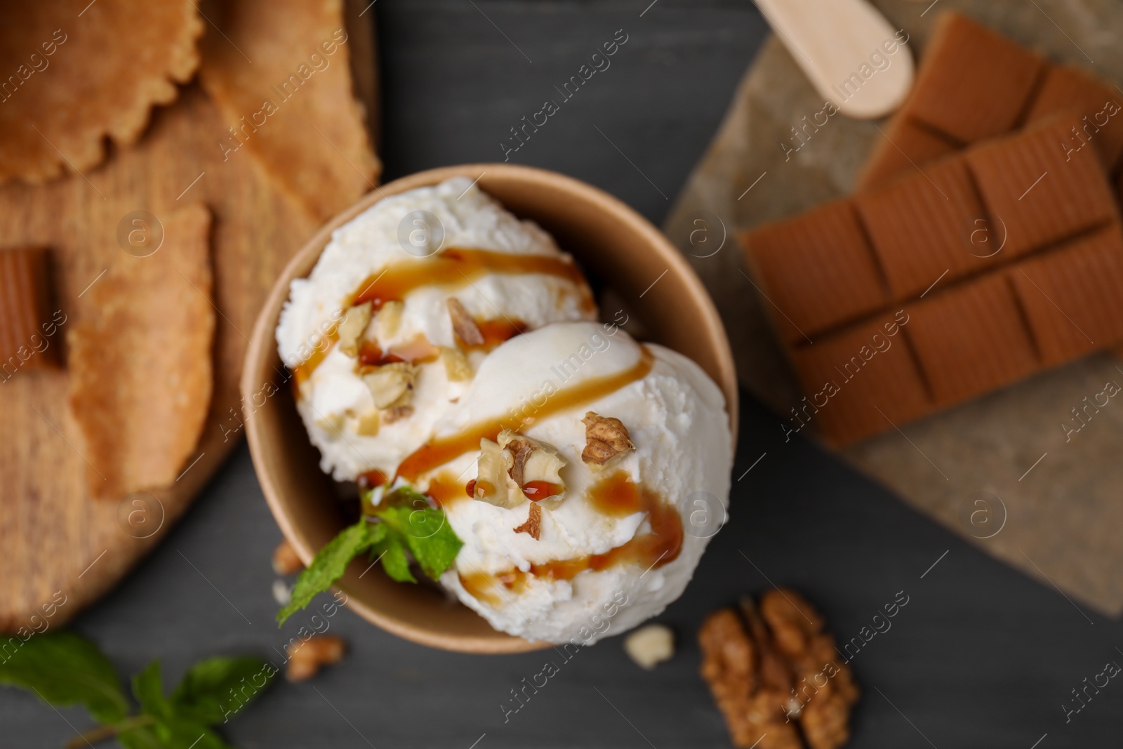 Photo of Tasty ice cream with caramel sauce, mint and nuts in paper cup on grey table, flat lay