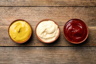 Photo of Ketchup, mustard and mayonnaise in bowls on wooden table, flat lay