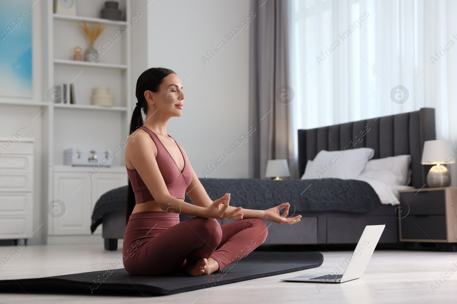 Photo of Beautiful young woman practicing Padmasana with laptop on yoga mat at home. Lotus pose