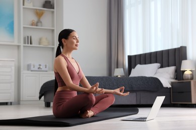 Photo of Beautiful young woman practicing Padmasana with laptop on yoga mat at home. Lotus pose