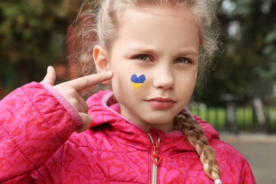 Photo of Little girl with drawing of Ukrainian flag on face in heart shape outdoors