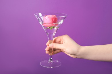 Photo of Woman holding beautiful martini glass with water and pink flower on purple background, closeup