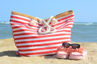 Stylish striped bag with slippers and sunglasses on sandy beach near sea