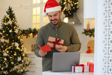 Celebrating Christmas online with exchanged by mail presents. Happy man in Santa hat with greeting card and gift box during video call on laptop in kitchen
