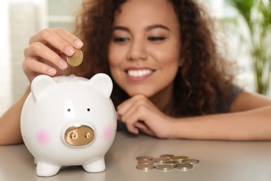 Photo of Happy African-American woman putting coin into piggy bank at table, focus on hand