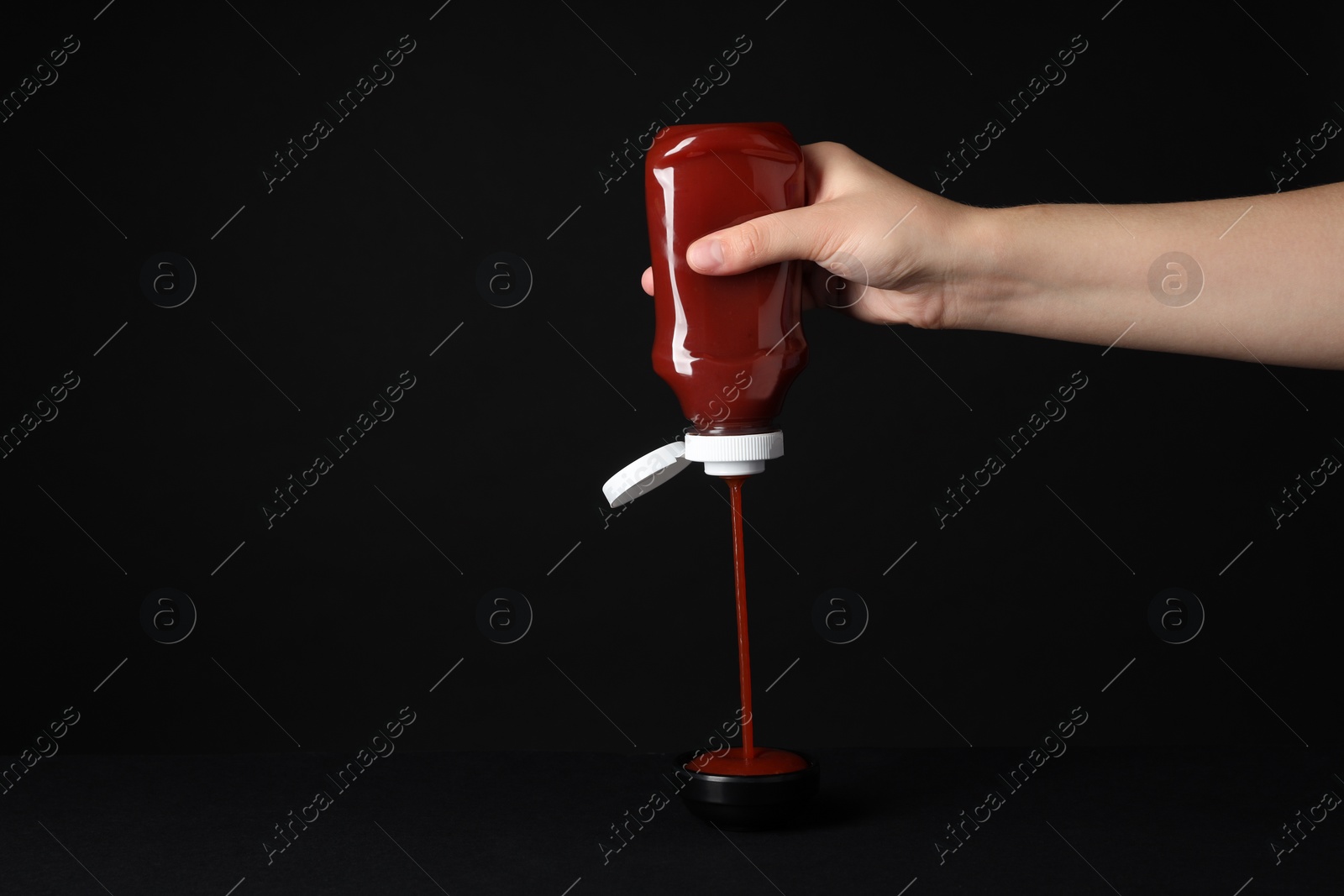 Photo of Woman pouring tasty ketchup from bottle into bowl on black background, closeup. Space for text