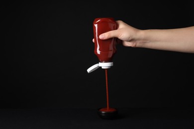 Woman pouring tasty ketchup from bottle into bowl on black background, closeup. Space for text