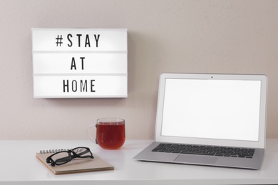 Photo of Laptop, cup of tea and lightbox with hashtag STAY AT HOME on white wall. Message to promote self-isolation during COVID‑19