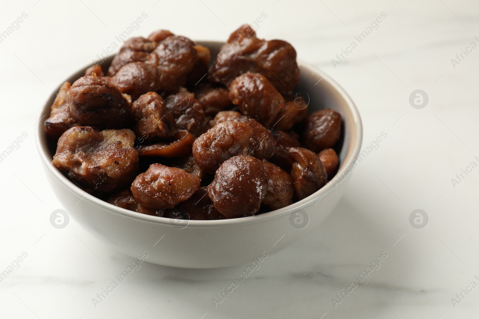 Photo of Roasted edible sweet chestnuts in bowl on white marble table, closeup