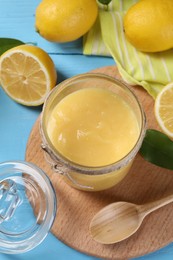 Photo of Delicious lemon curd in glass jar, fresh citrus fruits, green leaves and spoon on light blue wooden table