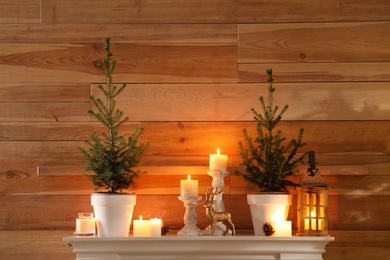 Photo of Small potted firs, candles and decor elements on white mantel near wooden wall
