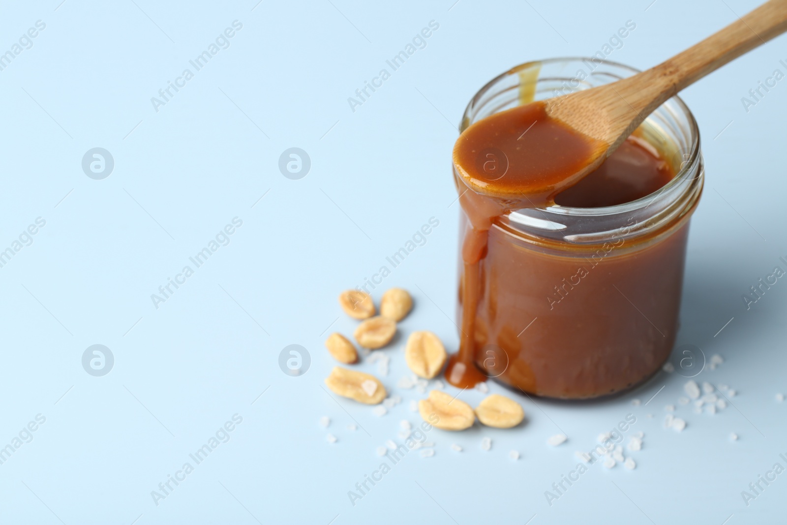 Photo of Jar of tasty salted caramel, spoon and peanuts on light blue background, closeup. Space for text