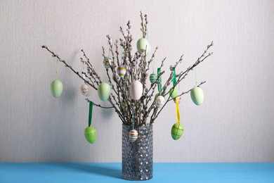 Photo of Beautiful willow branches with painted eggs in vase on light blue table. Easter decor