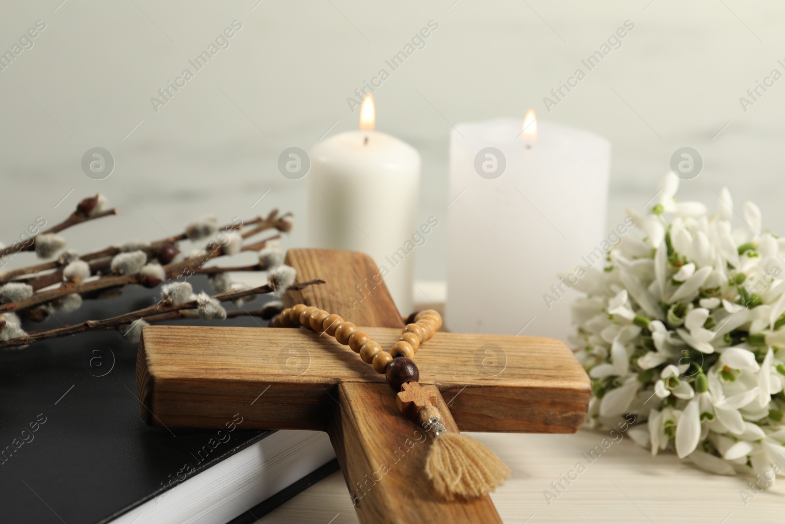 Photo of Burning church candles, wooden cross, rosary beads, Bible, willow branches and snowdrops on white table, closeup