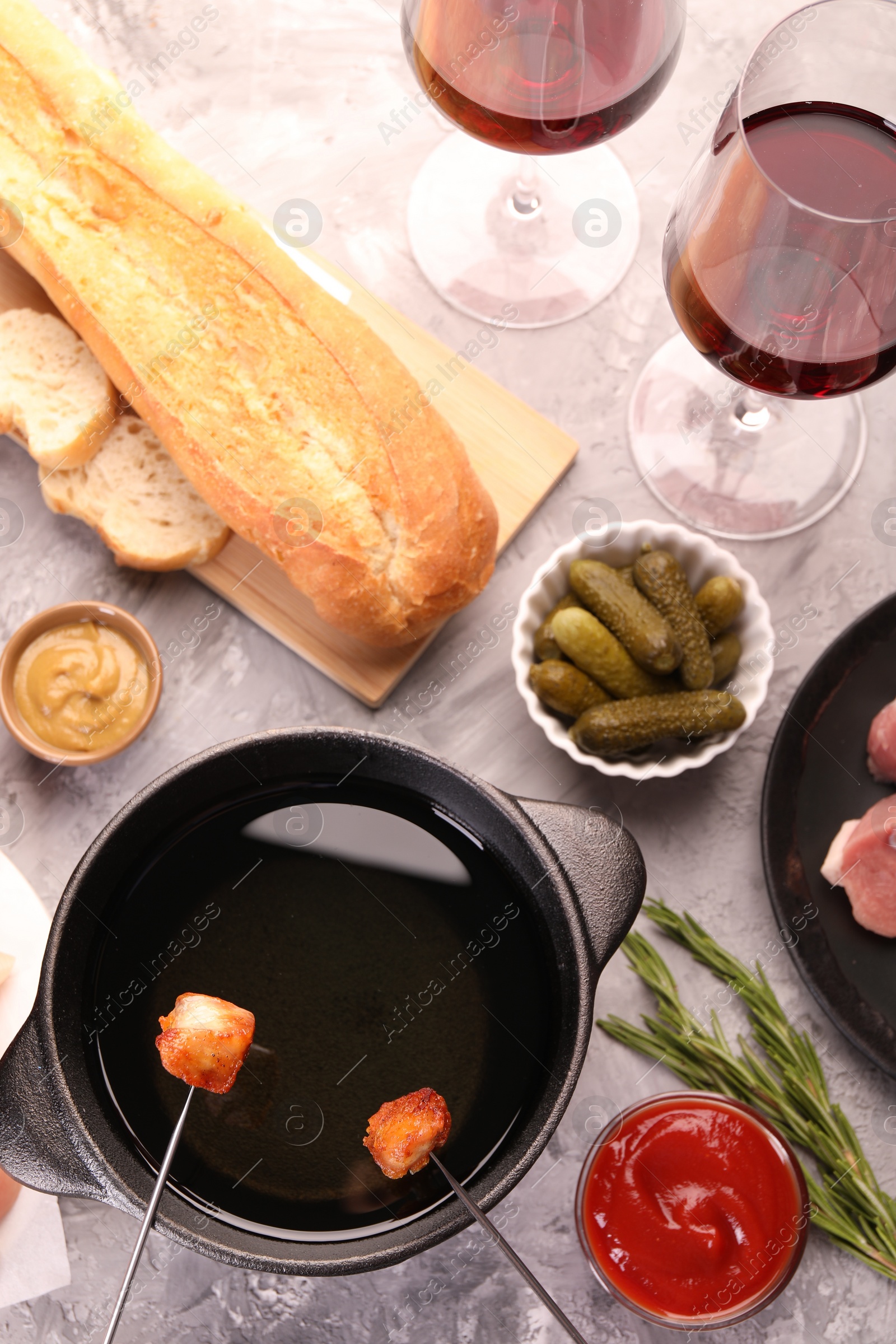 Photo of Fondue pot, forks with fried meat pieces, glasses of red wine and other products on grey textured table, flat lay
