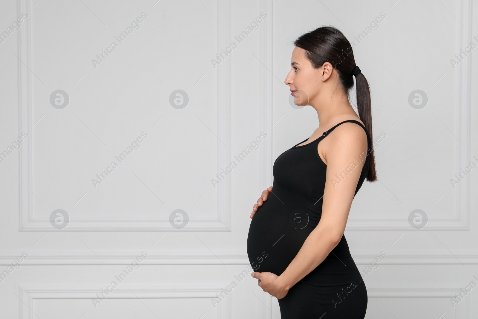 Photo of Beautiful pregnant woman in black dress near light grey wall, space for text