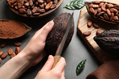 Photo of Woman cutting ripe cocoa pod over grey table with products, closeup