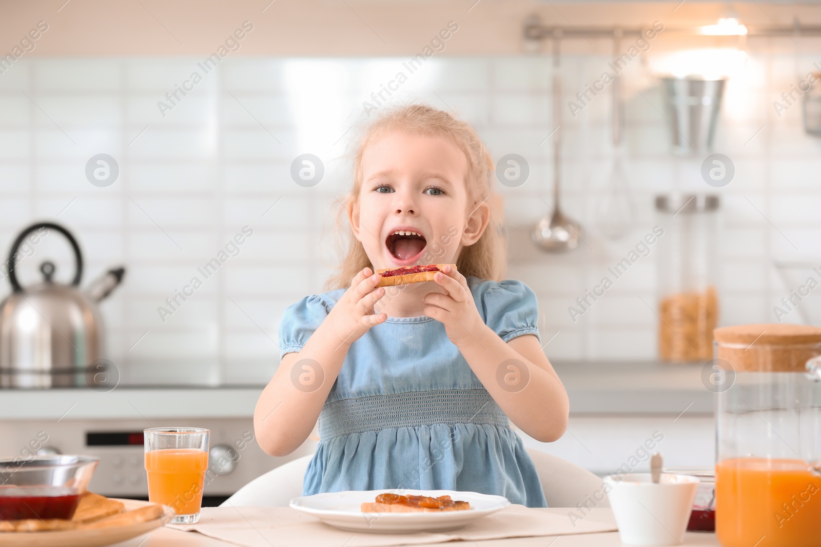 Photo of Adorable little girl eating tasty toasted bread with jam at table in kitchen