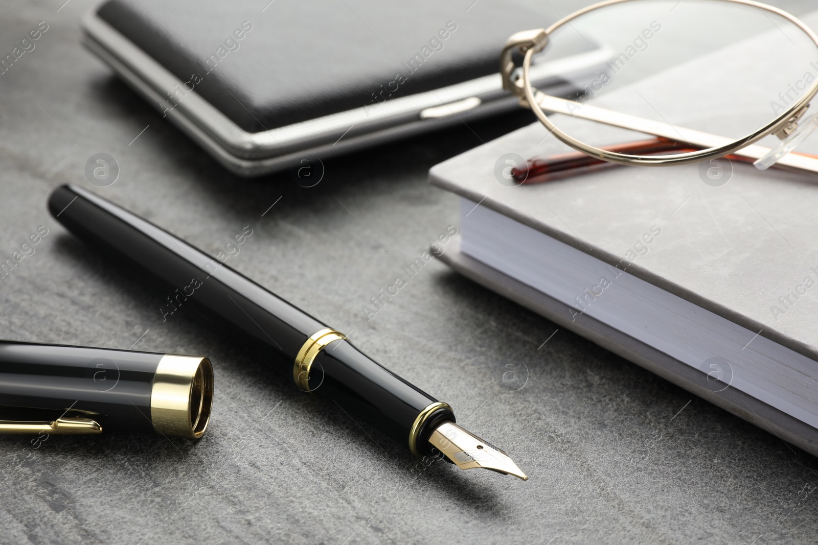 Photo of Stylish black fountain pen, glasses, notebook and cigarette case on grey textured table, closeup