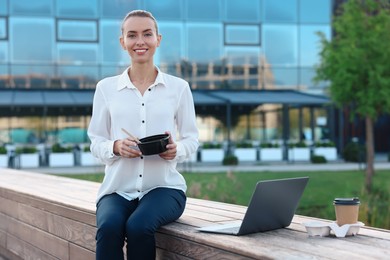 Photo of Smiling businesswoman with lunch box sitting near laptop and paper cup outdoors