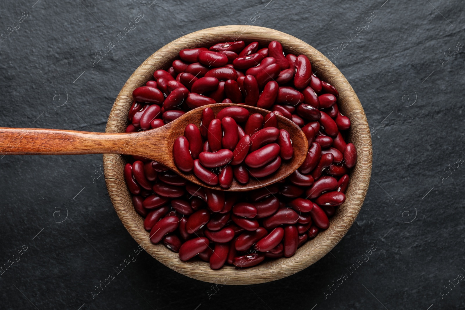 Photo of Raw red kidney beans with wooden spoon and bowl on black table, top view