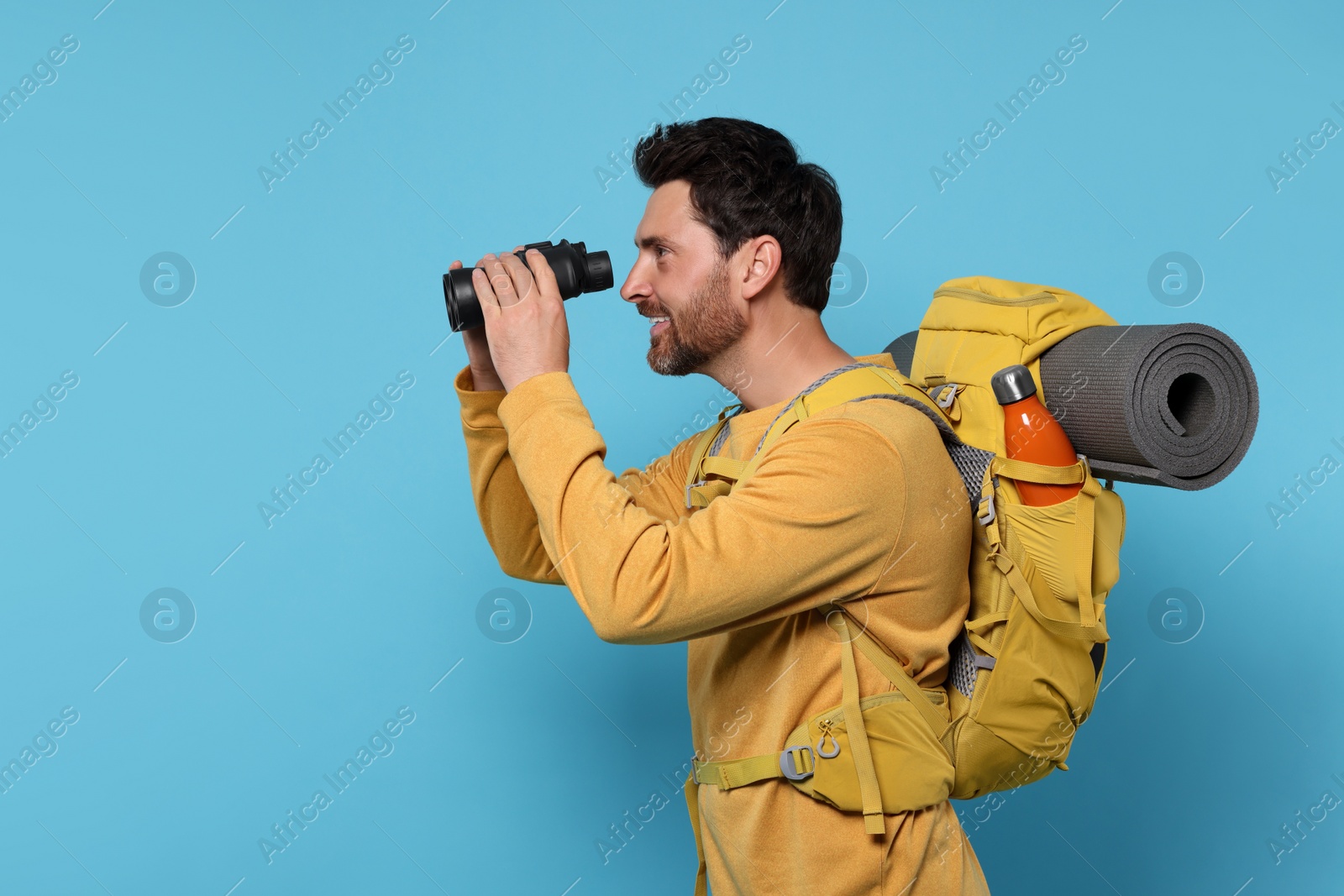 Photo of Happy man with backpack looking through binoculars on light blue background, space for text. Active tourism
