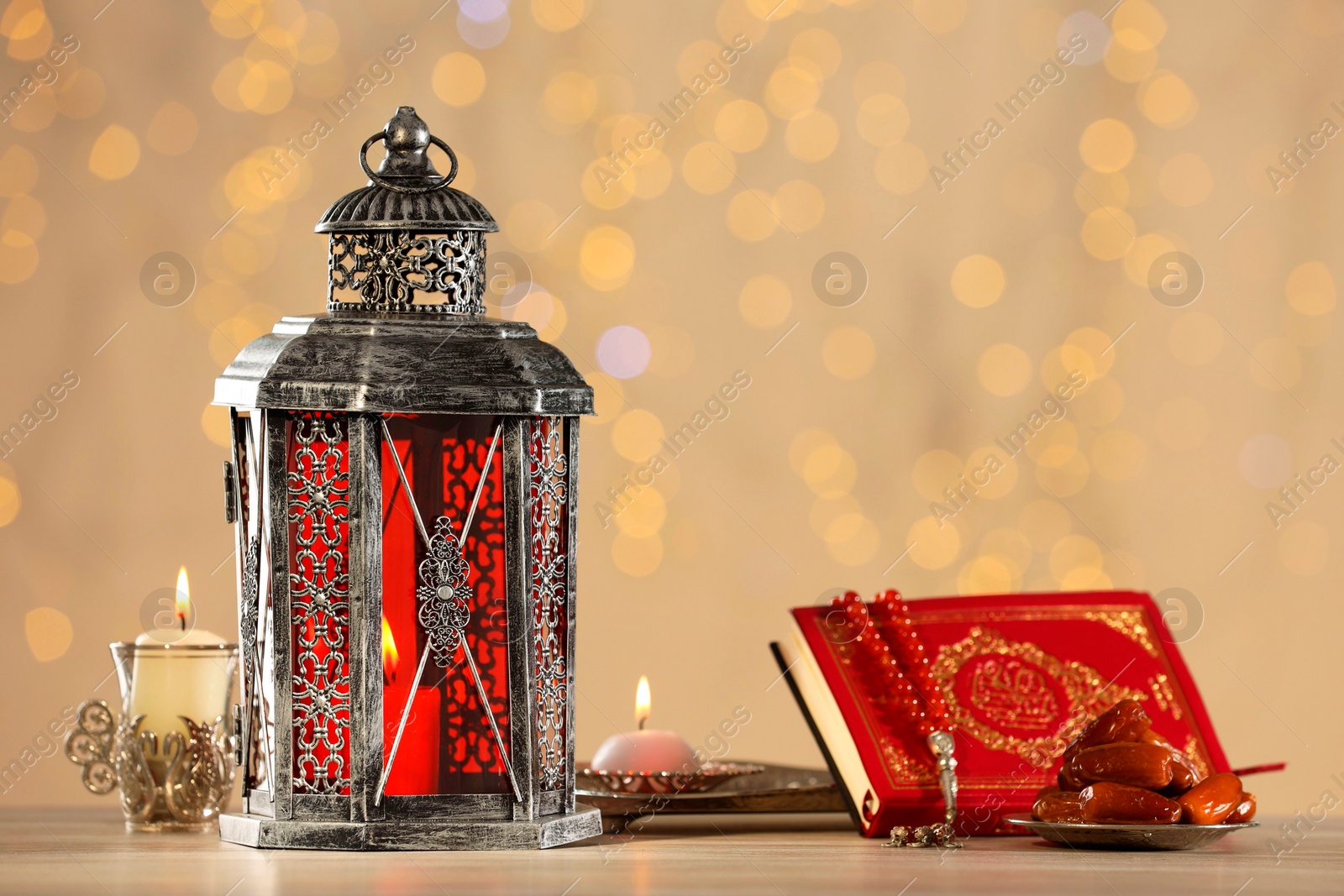 Photo of Arabic lantern, Quran, misbaha, candles and dates on wooden table against blurred lights