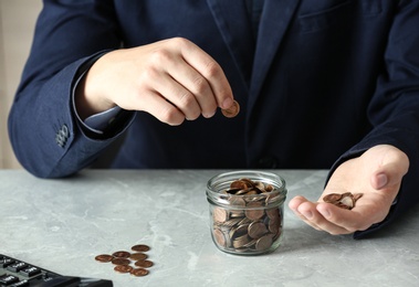Man putting coin into jar at grey marble table, closeup. Money savings