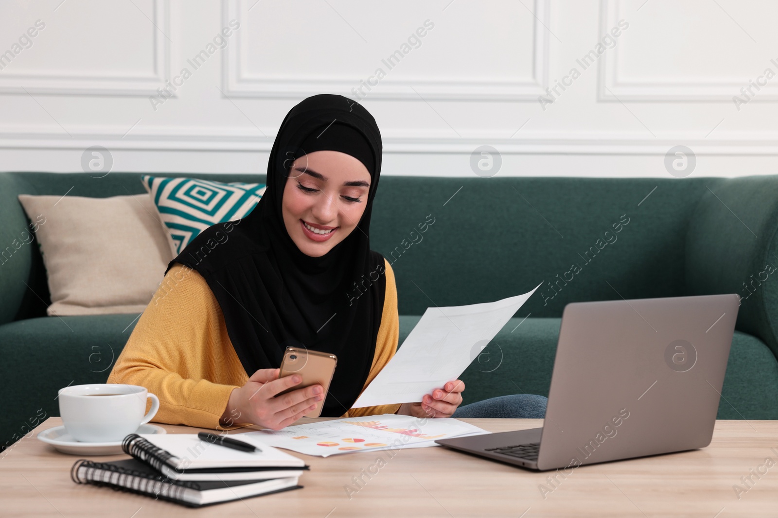 Photo of Muslim woman in hijab using laptop and smartphone at wooden table indoors