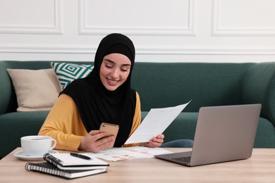 Muslim woman in hijab using laptop and smartphone at wooden table indoors
