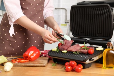 Woman cooking different products with electric grill at wooden table in kitchen, closeup