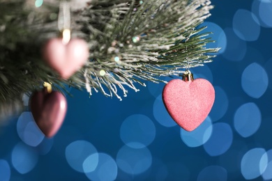 Photo of Beautiful holiday heart shaped bauble hanging on Christmas tree against blue background with blurred festive lights